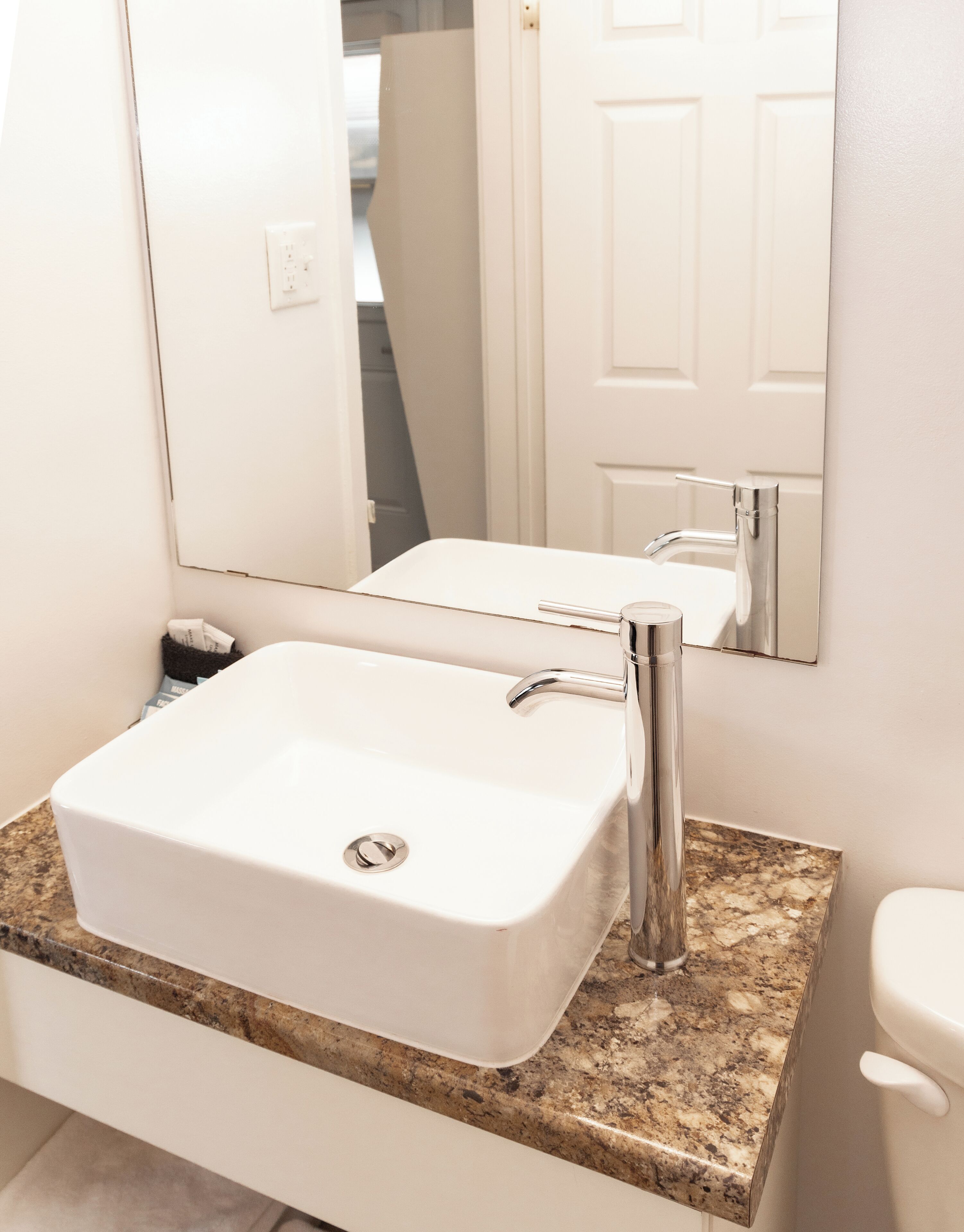 Un baño con lavamanos blanco, espejo, encimera de granito y retrete blanco.