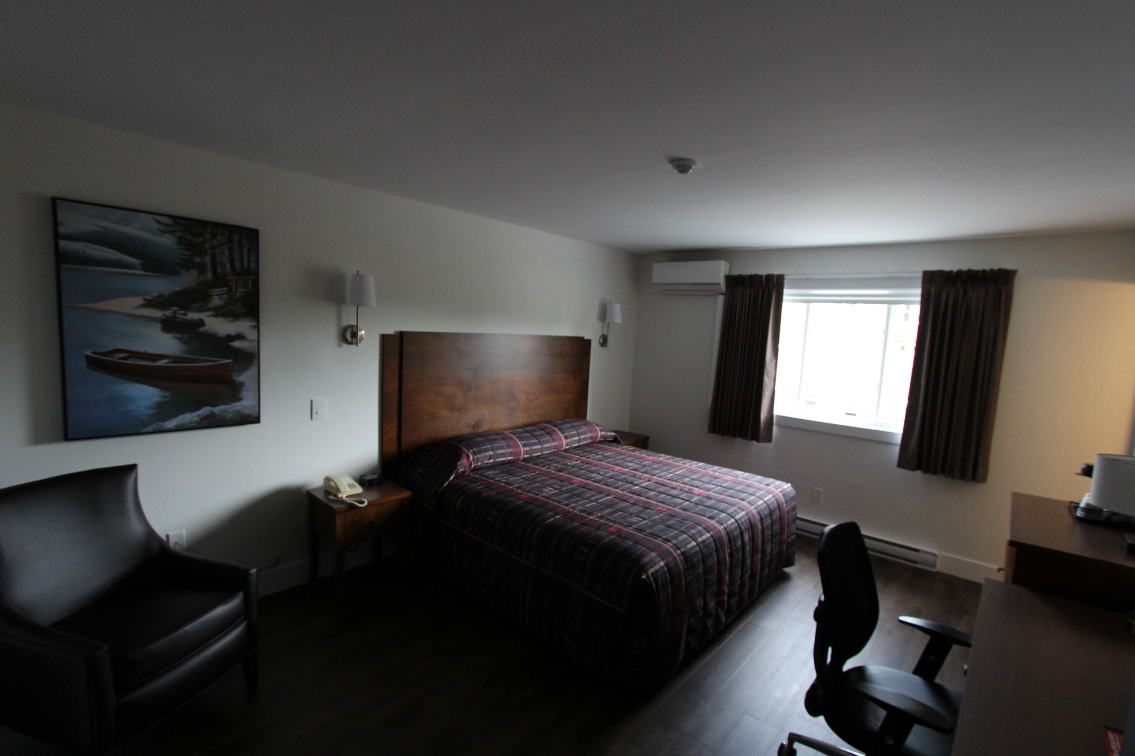A hotel room with a bed, a desk, a chair, a nightstand, a lamp, a painting, and a window with curtains.