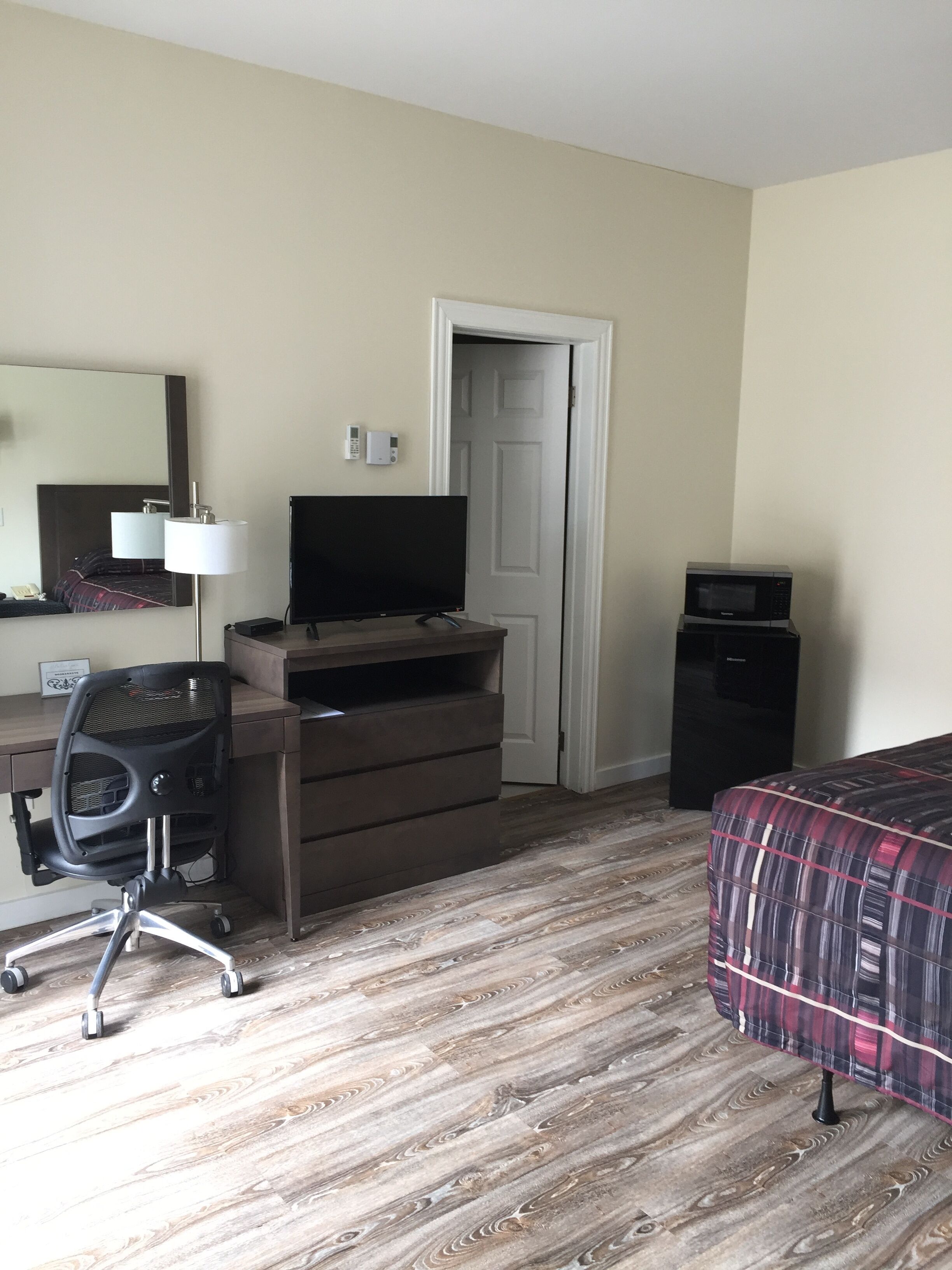 Una habitación de hotel con piso de madera, cama con cubrecama a cuadros, escritorio con computadora y lámpara, televisor, minibar y una puerta.