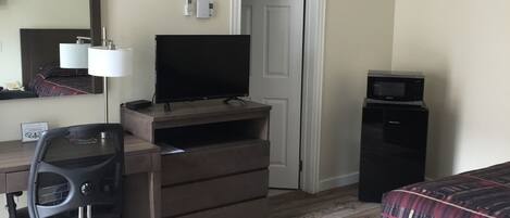 Una habitación de hotel con piso de madera, cama con cubrecama a cuadros, escritorio con computadora y lámpara, televisor, minibar y una puerta.