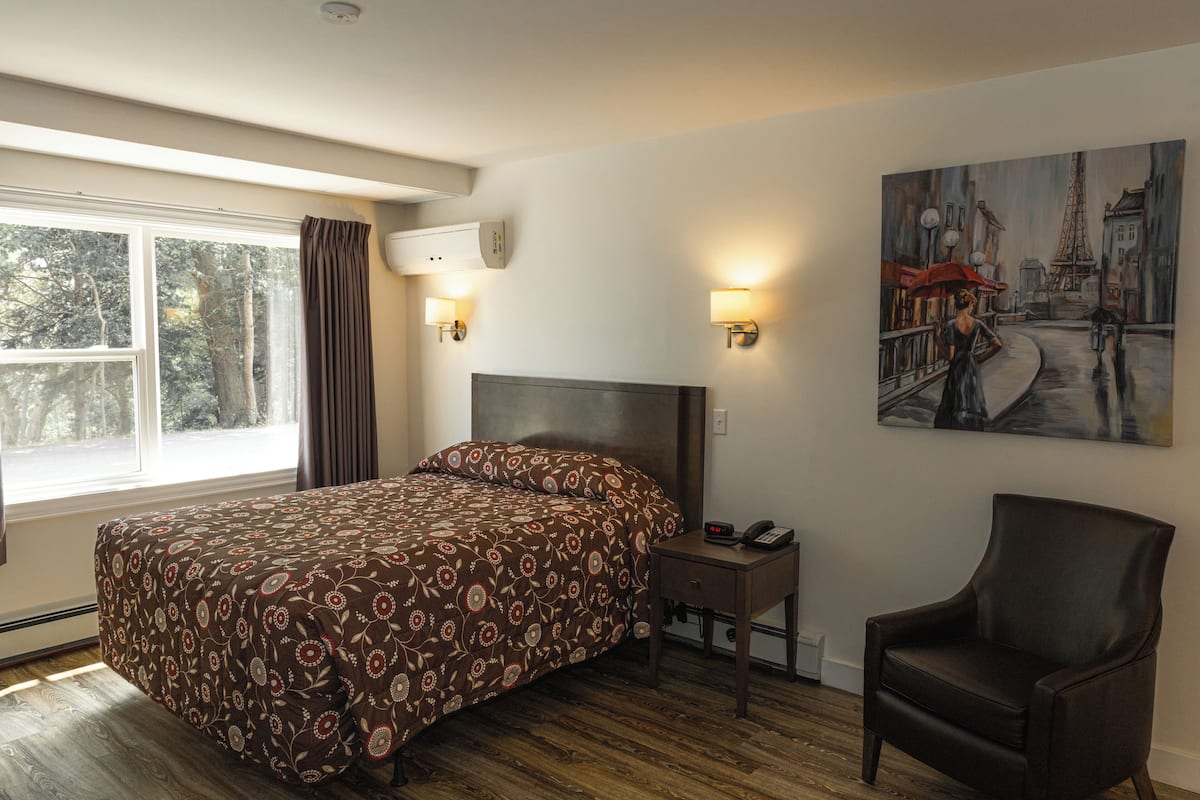A hotel room with a large bed, bedside table with a phone, a chair, a painting on the wall, and a window with curtains.