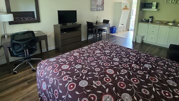 A hotel room with a large bed, a desk with a chair, a television, a small table with a plant, a picture on the wall, and a kitchenette area.