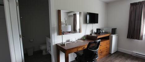 Ein Zimmer mit Schreibtisch aus Holz, schwarzem Bürostuhl, Wand-Fernseher, kleinem Kühlschrank und einem Fenster mit Vorhängen.