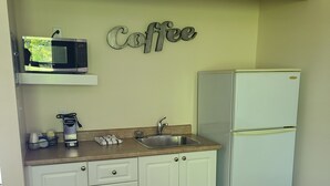 Une cuisine compacte avec des meubles blancs, un évier en acier inoxydable, un micro-ondes posé sur une étagère, un réfrigérateur et un panneau indiquant « Café » sur le mur.