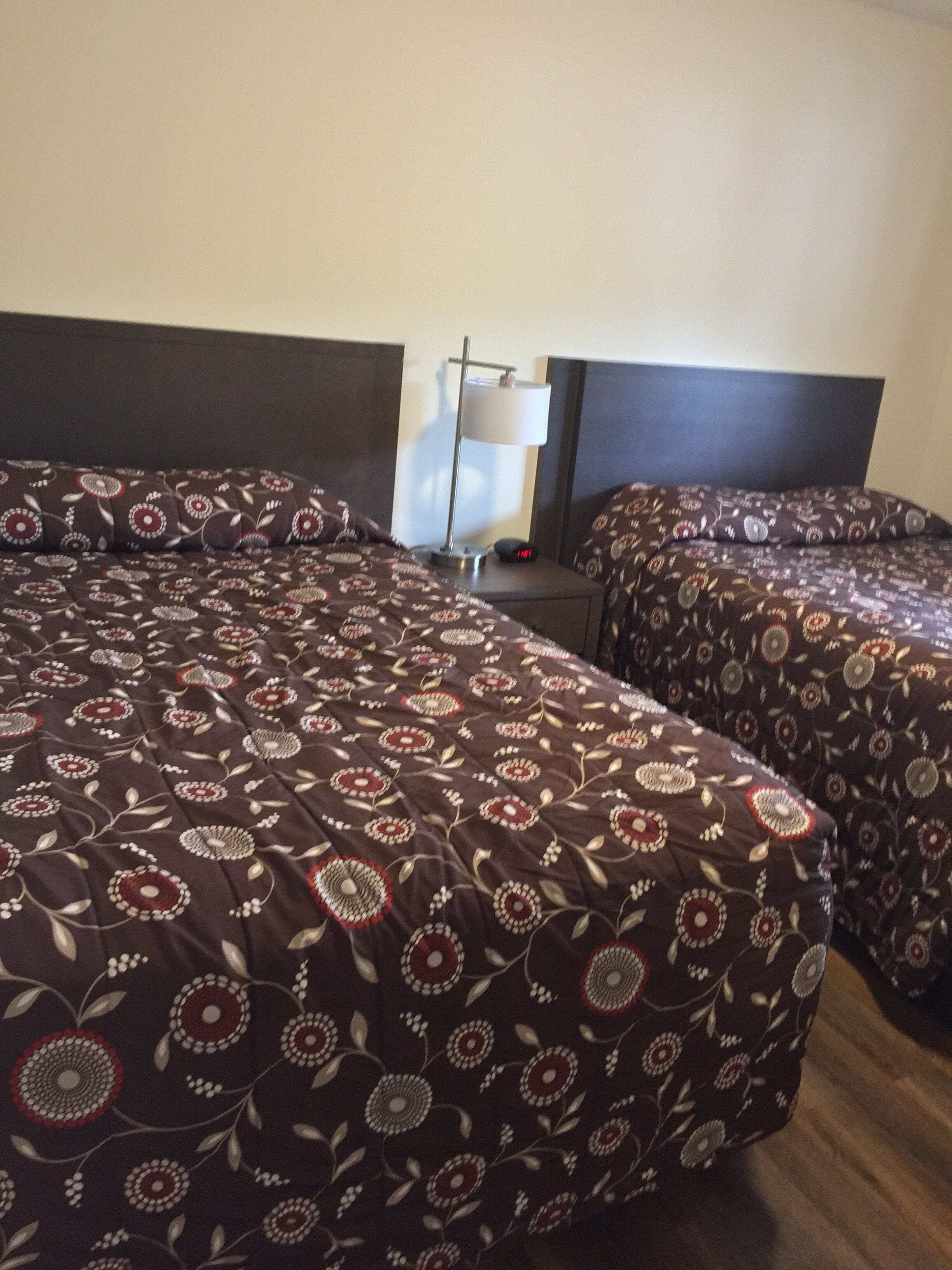 Habitación de hotel con dos camas, cada una con cubrecama estampado con motivos florales. Se observa una mesita de noche con una lámpara y un reloj.