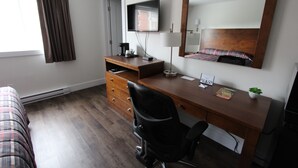Une chambre d’hôtel avec un bureau en bois, un fauteuil de bureau noir, un téléviseur à écran plat, un miroir, une table de chevet, une lampe, une petite plante en pot et une couvre-lit à motifs écossais.