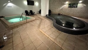 An indoor swimming pool with a jacuzzi, tiled floor, and black lounge chairs.