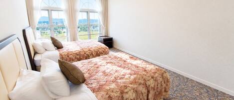 A hotel room with two beds, a nightstand, and a view of a cityscape through large windows.