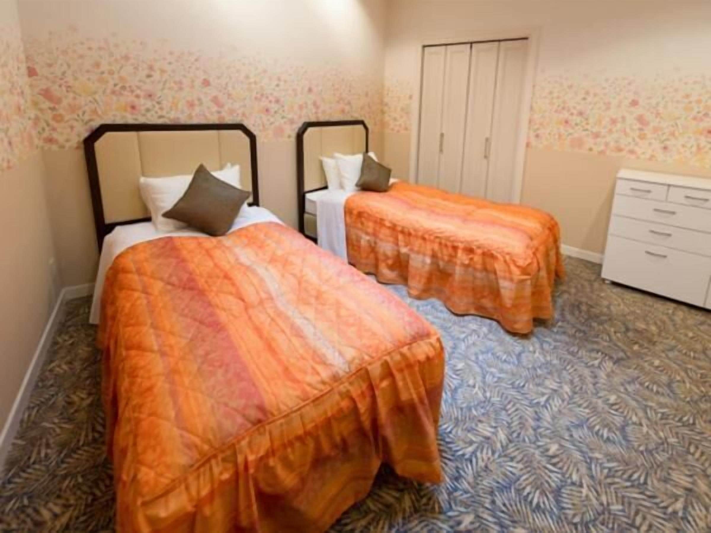A room with two beds, each with an orange bedspread and white pillows. There is a wardrobe with a mirror and a small table with a lamp.