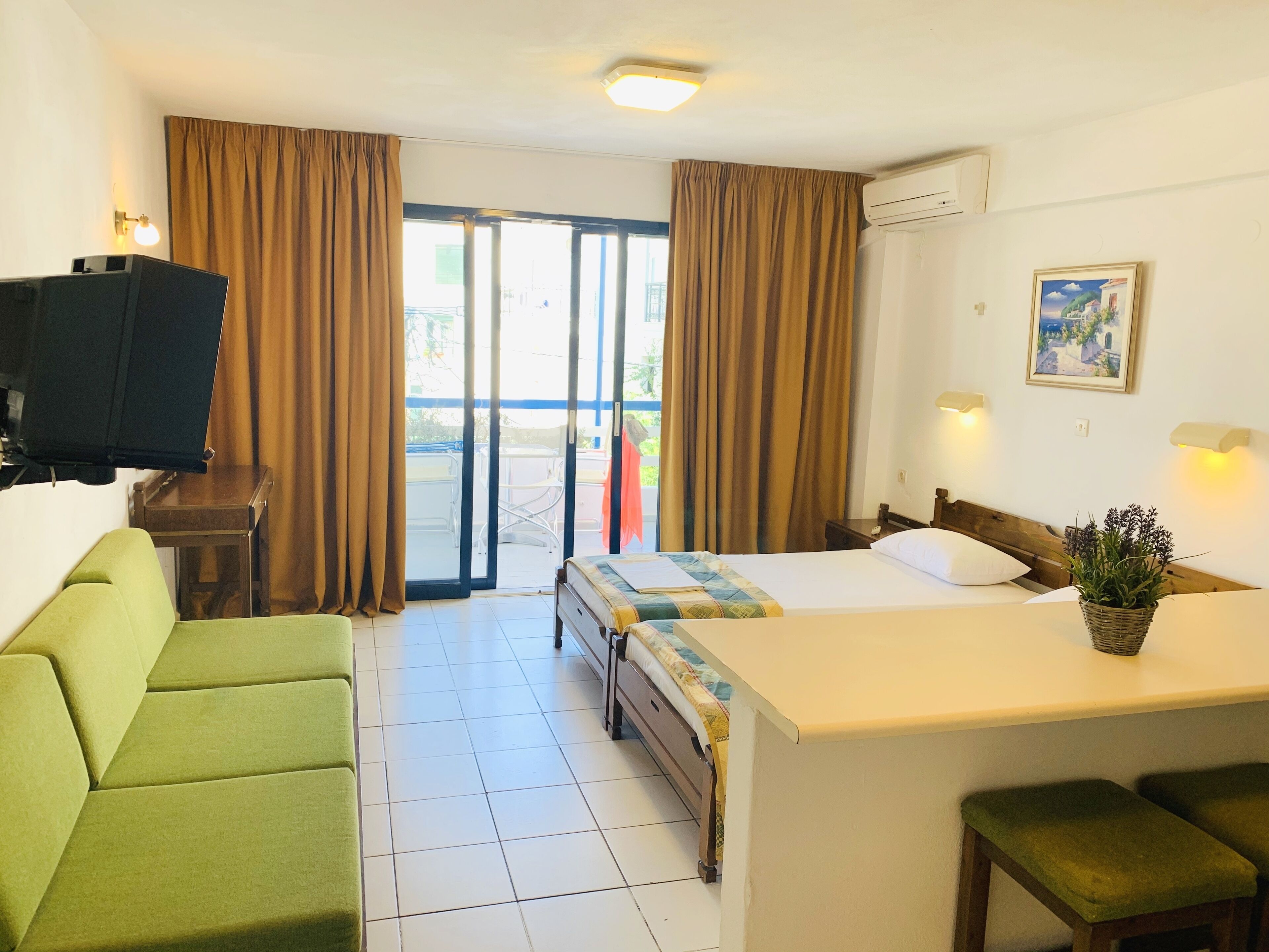 A hotel room with a bed, a small desk, a television, a green sofa, and a view through a sliding door to the outside.
