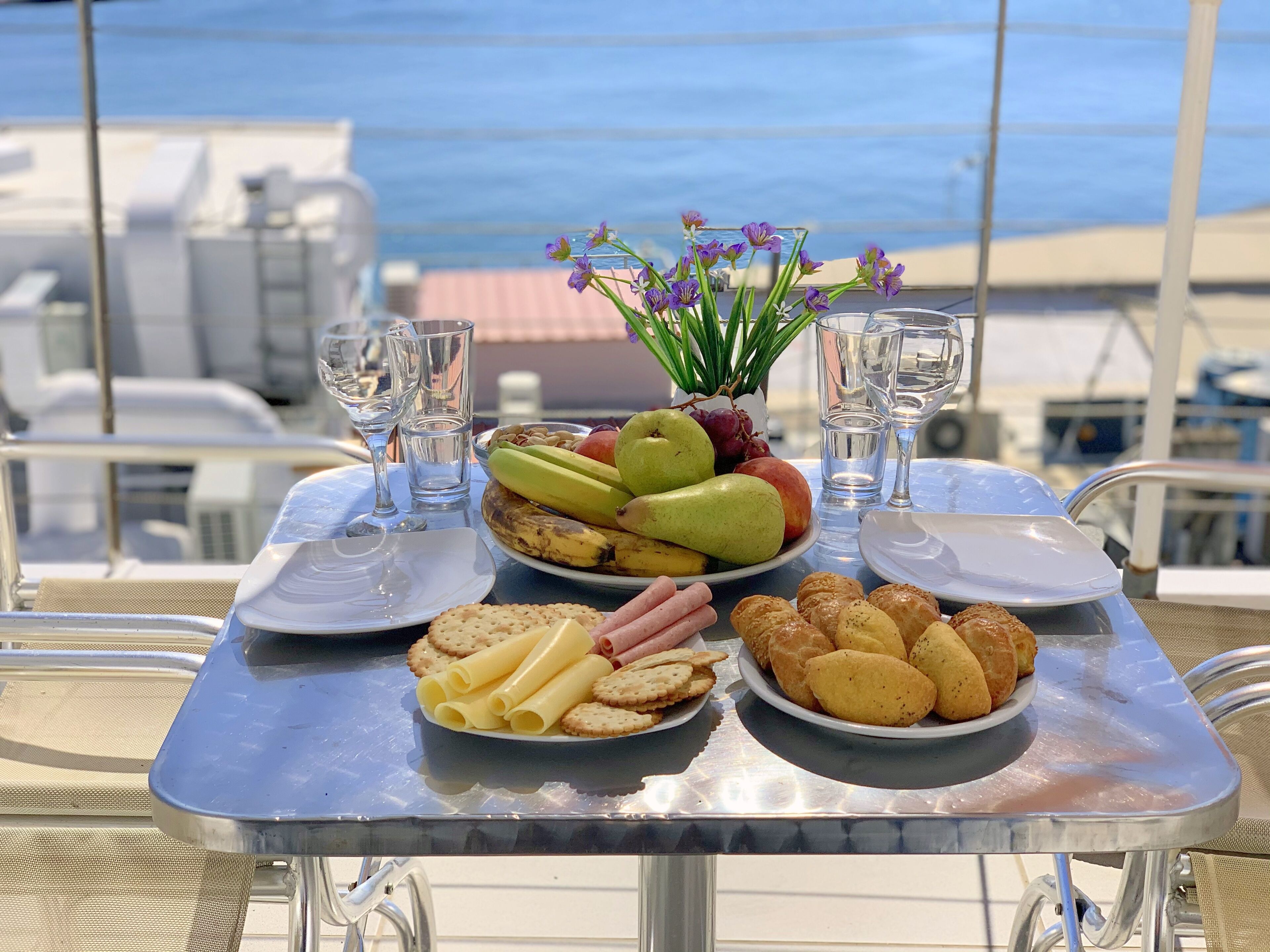 Una mesa con una bandeja de frutas, galletas y pasteles, vasos y un jarrón de flores en un balcón con vistas al mar.