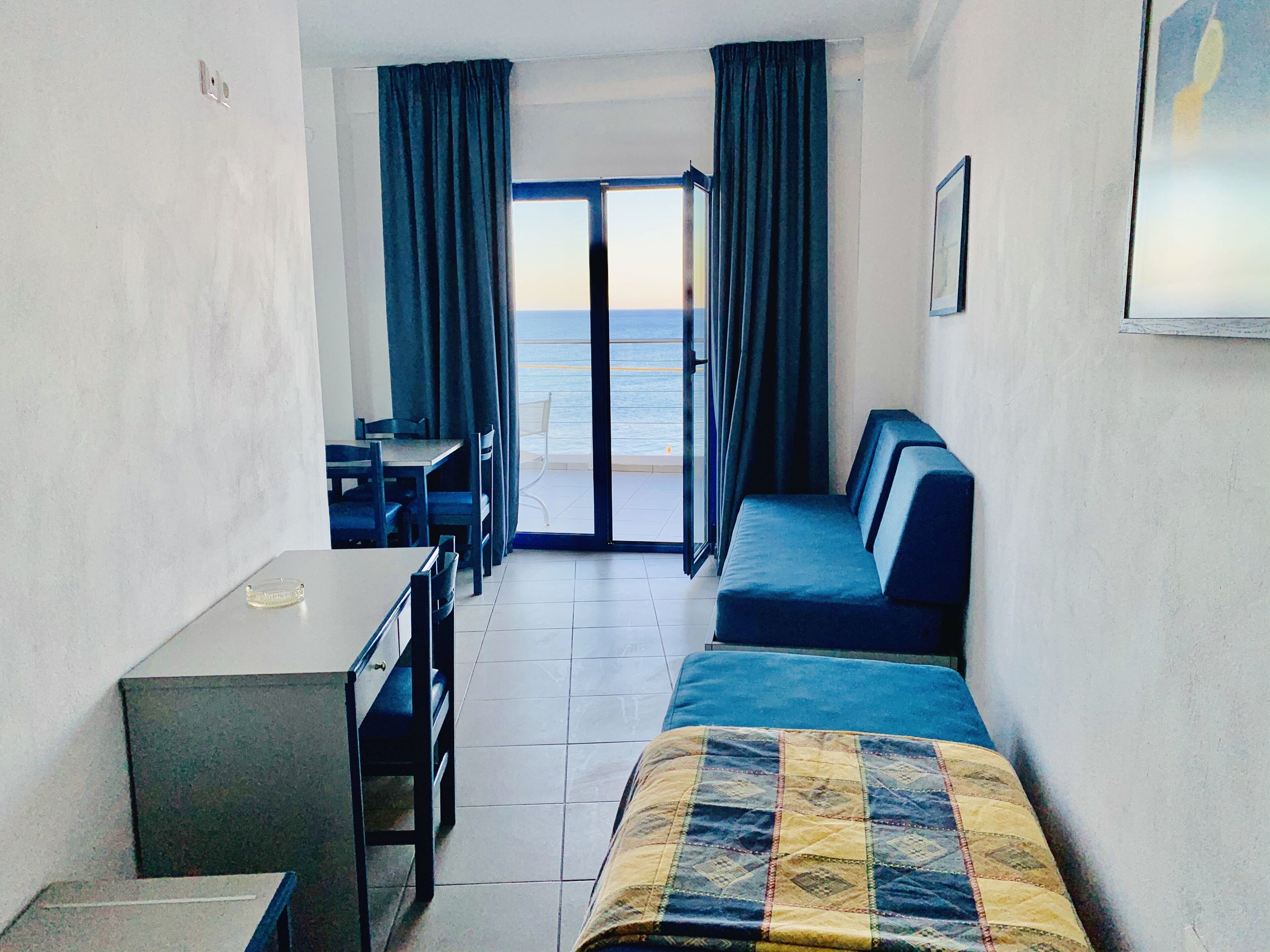 A room with a bed, a table, and a chair. There is a view of the ocean through the window.