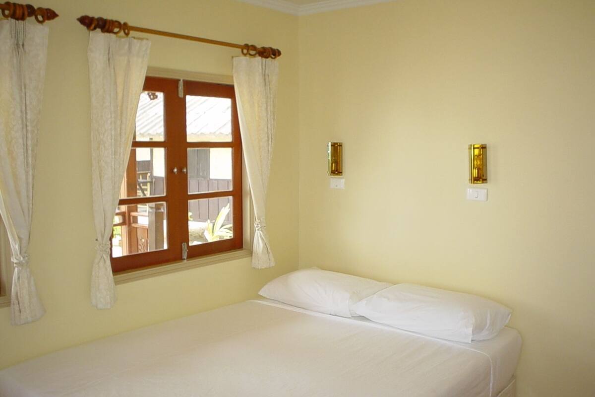 Una cama individual con sábanas blancas en una habitación con paredes color crema, una puerta de madera y una ventana con cortinas.