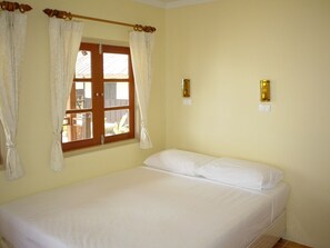 A single bed with white linens in a room with cream walls, a wooden door, and a window with curtains.