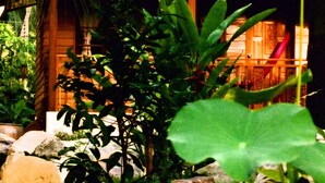 Una casa di legno con tetto di paglia, circondata da vegetazione rigogliosa e uno stagno con rocce e piante.