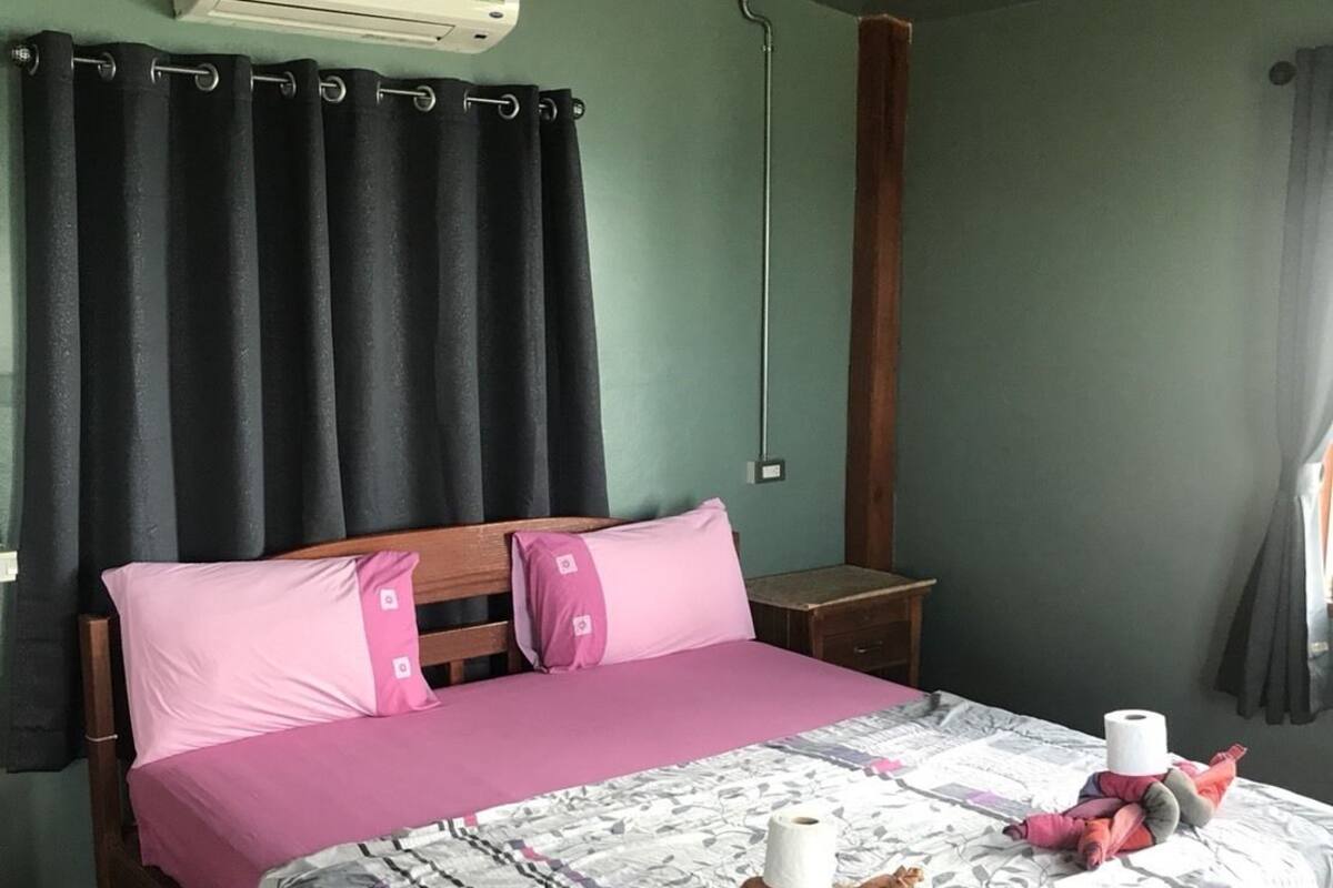 A bedroom with a bed, a wooden nightstand, a wall-mounted air conditioner, and a window with curtains.