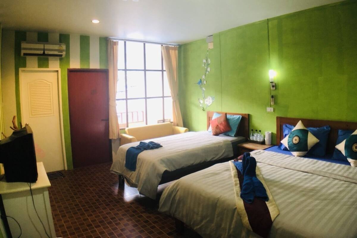 A hotel room with two beds, a window with curtains, a desk with a computer, and a green wall with a decorative branch.
