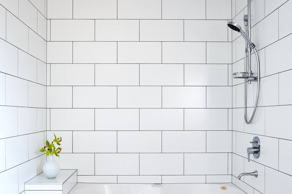 A modern bathroom with white tiled walls, a white bathtub, and a small white vase with flowers on a pedestal.