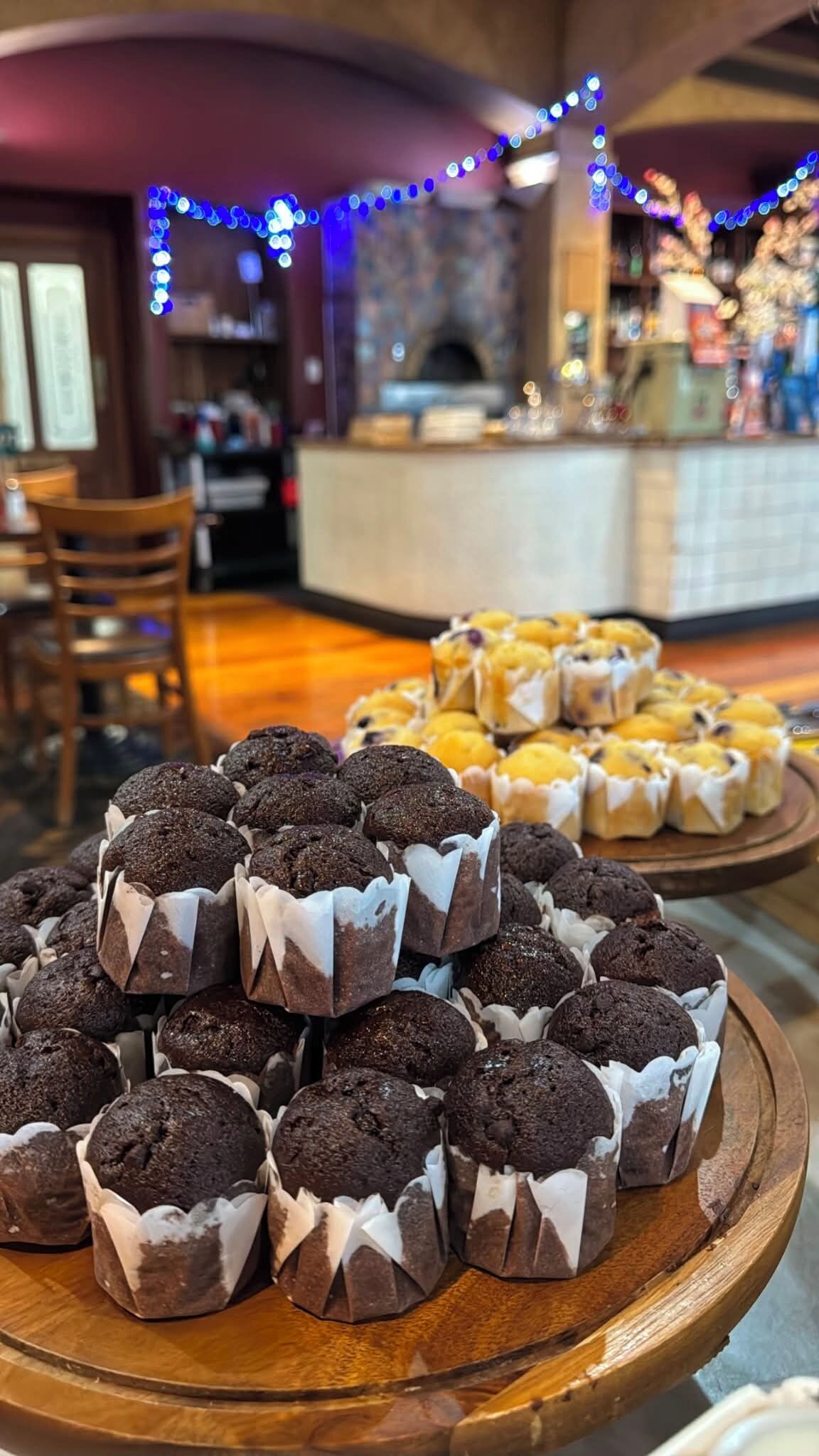 Variedad de productos horneados, incluyendo opciones de chocolate y masa de huevo, expuestos en bandejas de madera en un ambiente interior acogedor, con una barra de bar al fondo.