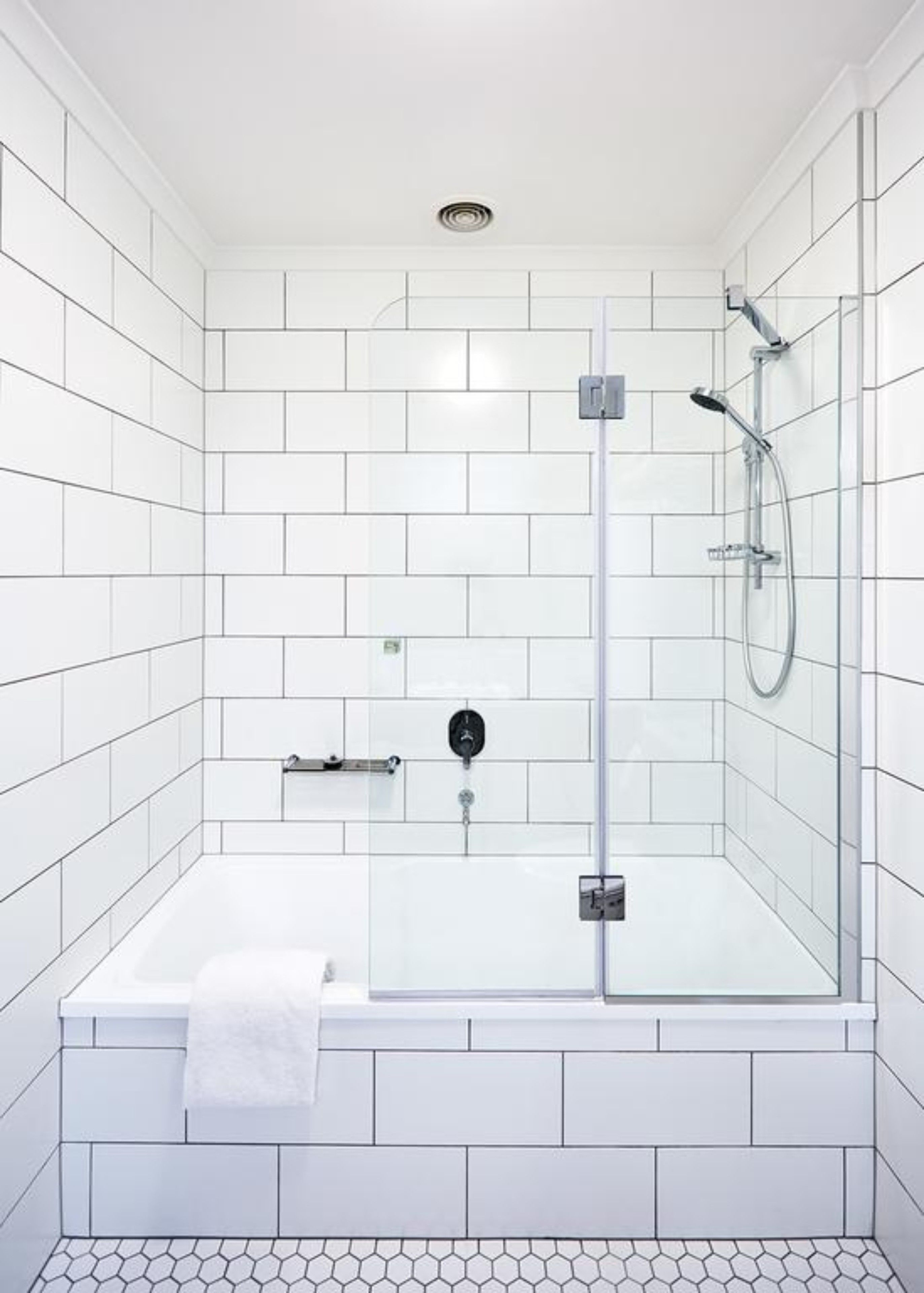 Un baño moderno con paredes y suelo de baldosas blancas, cabina de ducha con mampara de vidrio y una toalla blanca en el suelo.