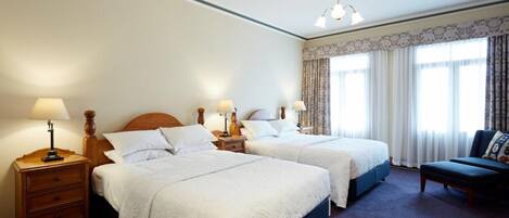 Habitación de hotel con dos camas, una mesita de noche con lámpara, una silla y un ventanal con cortinas.