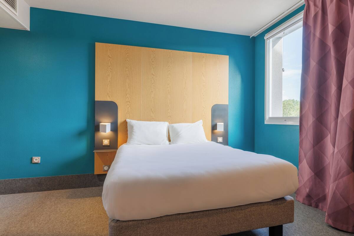 A hotel room with a large bed, wooden headboard, two bedside lamps, and a window with curtains.