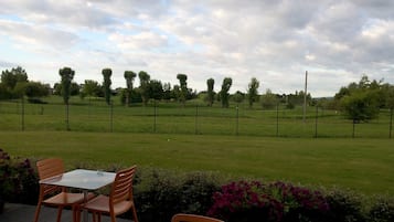 Uma área externa com mesas e cadeiras, cercada por um campo gramado e árvores, sob um céu nublado.