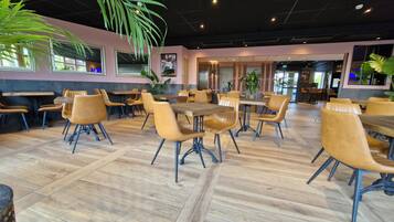 A spacious indoor dining area with wooden tables and chairs, large windows, and potted plants.