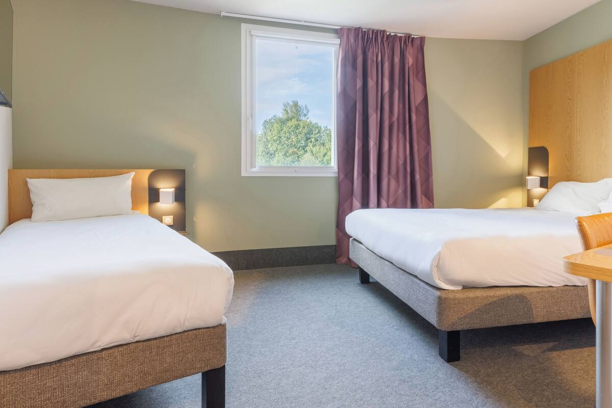 A hotel room with two beds, a window with curtains, and wall-mounted lamps.