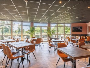 A spacious indoor dining area with large windows, wooden tables, and orange chairs. - B&B HOTEL Clermont-Ferand Nord Riom (Riom)