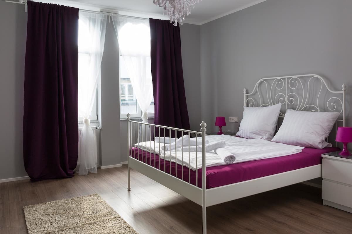 A bedroom with a white metal bed frame, a chandelier, a white nightstand, and purple curtains.