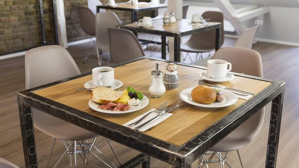 A breakfast setup with two plates of food, coffee cups, and a salt and pepper set on a wooden table in a modern dining area with high ceilings and brick walls. - Hotel Forty Four (Frankfurt)