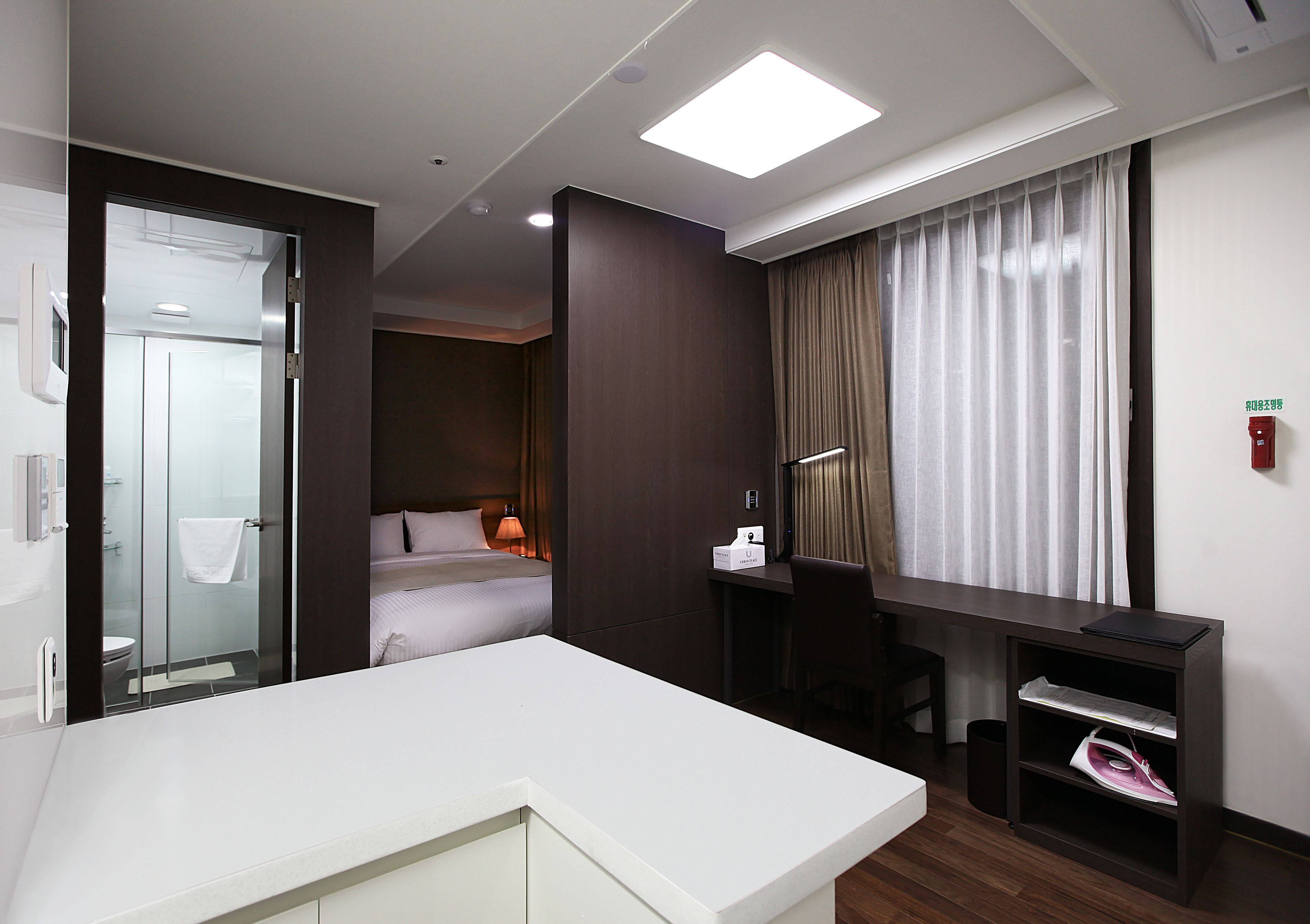 A modern hotel room with a white desk, a bed, a shower area, and a view of the city.