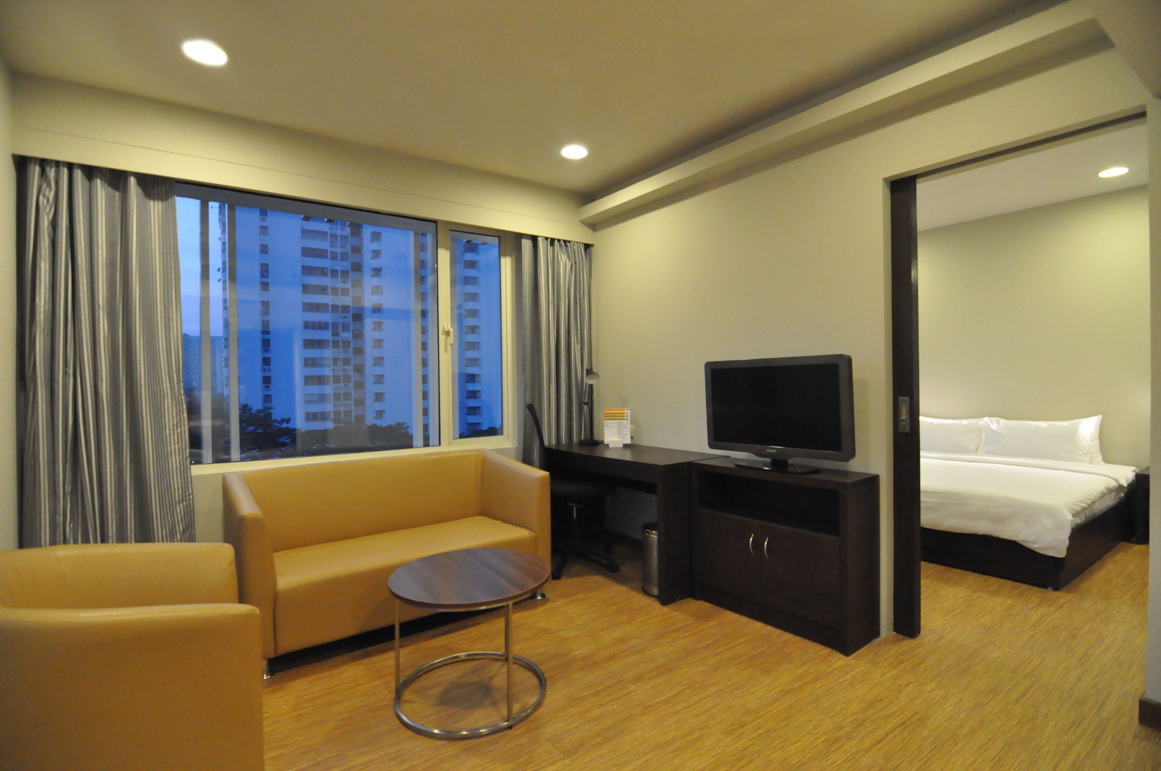 A hotel room with a sofa, a small table, a TV, a bed, and a view of buildings through the window.