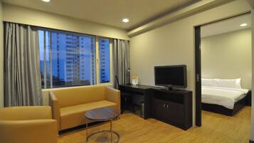 A hotel room with a sofa, a small table, a TV, a bed, and a view of buildings through the window.