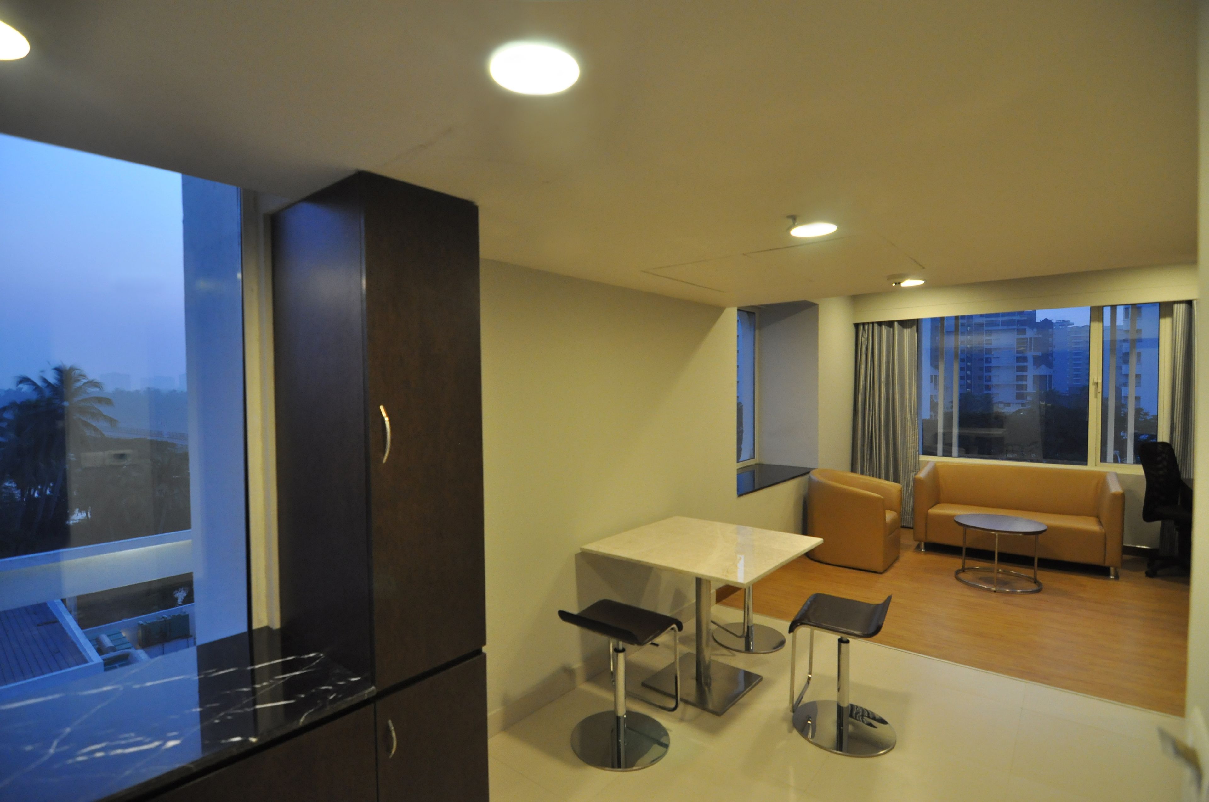 A modern hotel room with a dining area, a sofa, and a large window offering a city view.