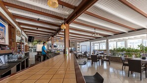 A spacious bar area with a long counter, wooden beams on the ceiling, and a view of the outdoor patio area with tables and chairs. - Mammis Beach (Kos)