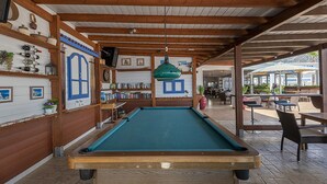 A pool table in a covered area with wooden beams, a bookshelf with books and decorative items, and a view of an outdoor seating area with tables and chairs. - Mammis Beach (Kos)