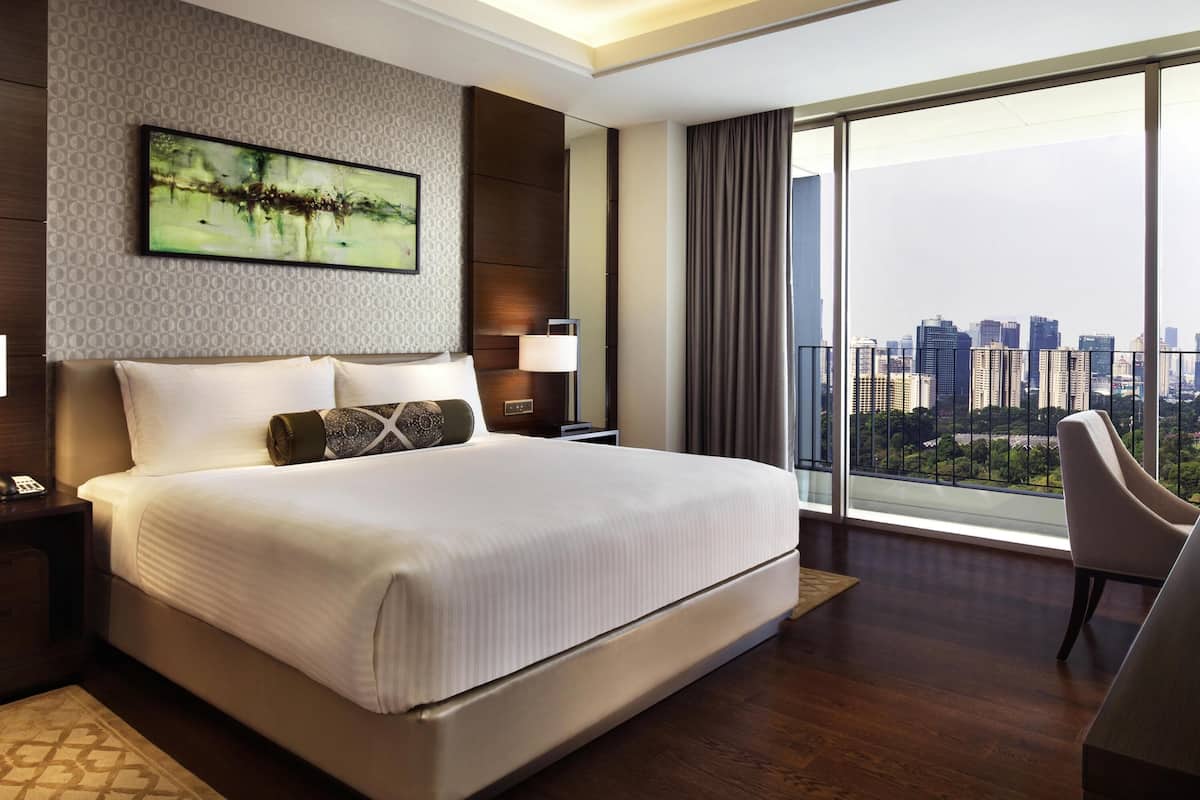 A modern hotel room with a large bed, a city view, a painting on the wall, and a balcony with a chair.
