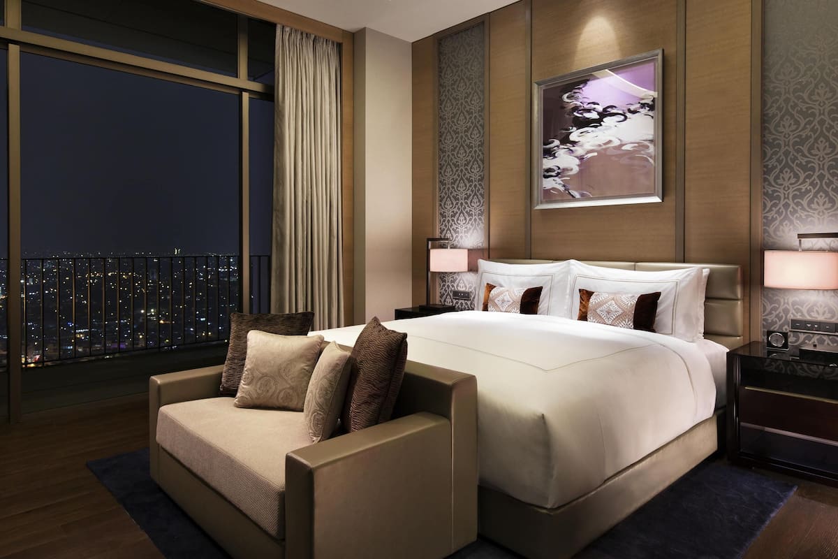 A modern hotel room with a large bed, a sofa, a city view, and a nightstand with a lamp.