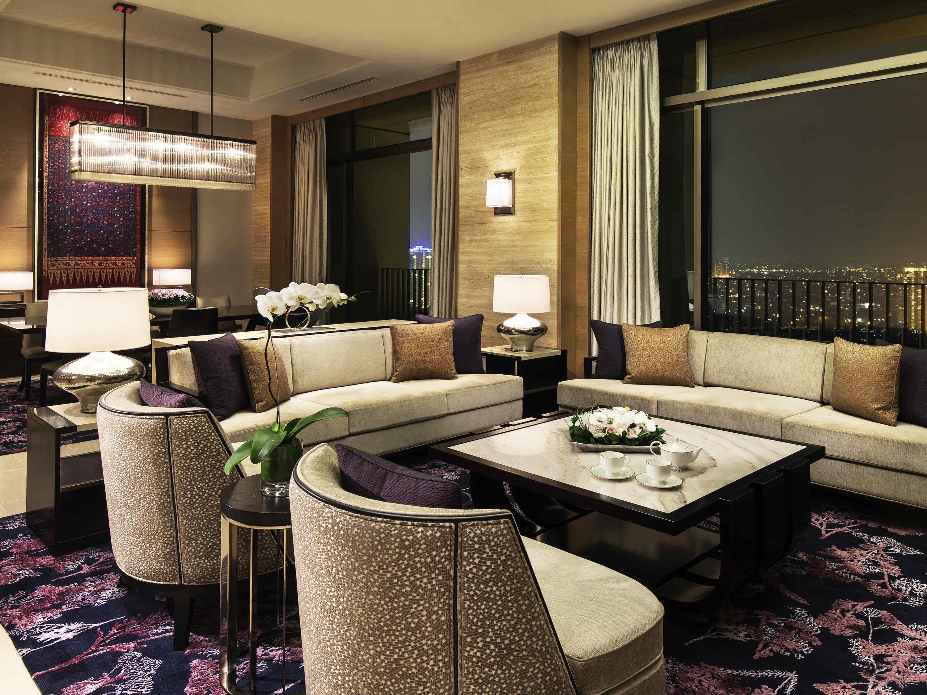Een moderne hotelkamer met een hoekbank, een ronde salontafel, een bijzettafeltje met een lamp en een uitzicht op de stad door grote ramen.