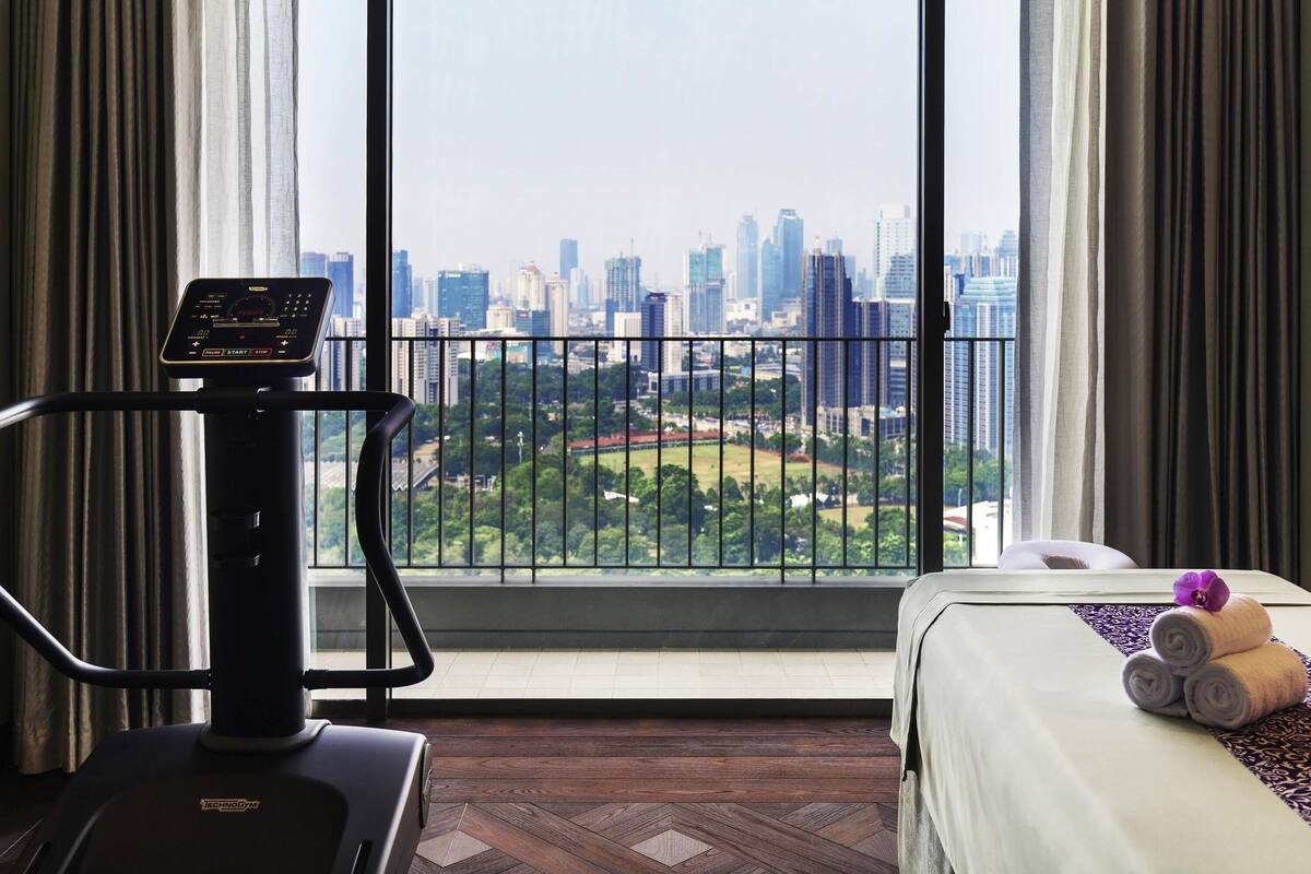 A hotel room with a treadmill, a bed with towels, and a city view through large windows.