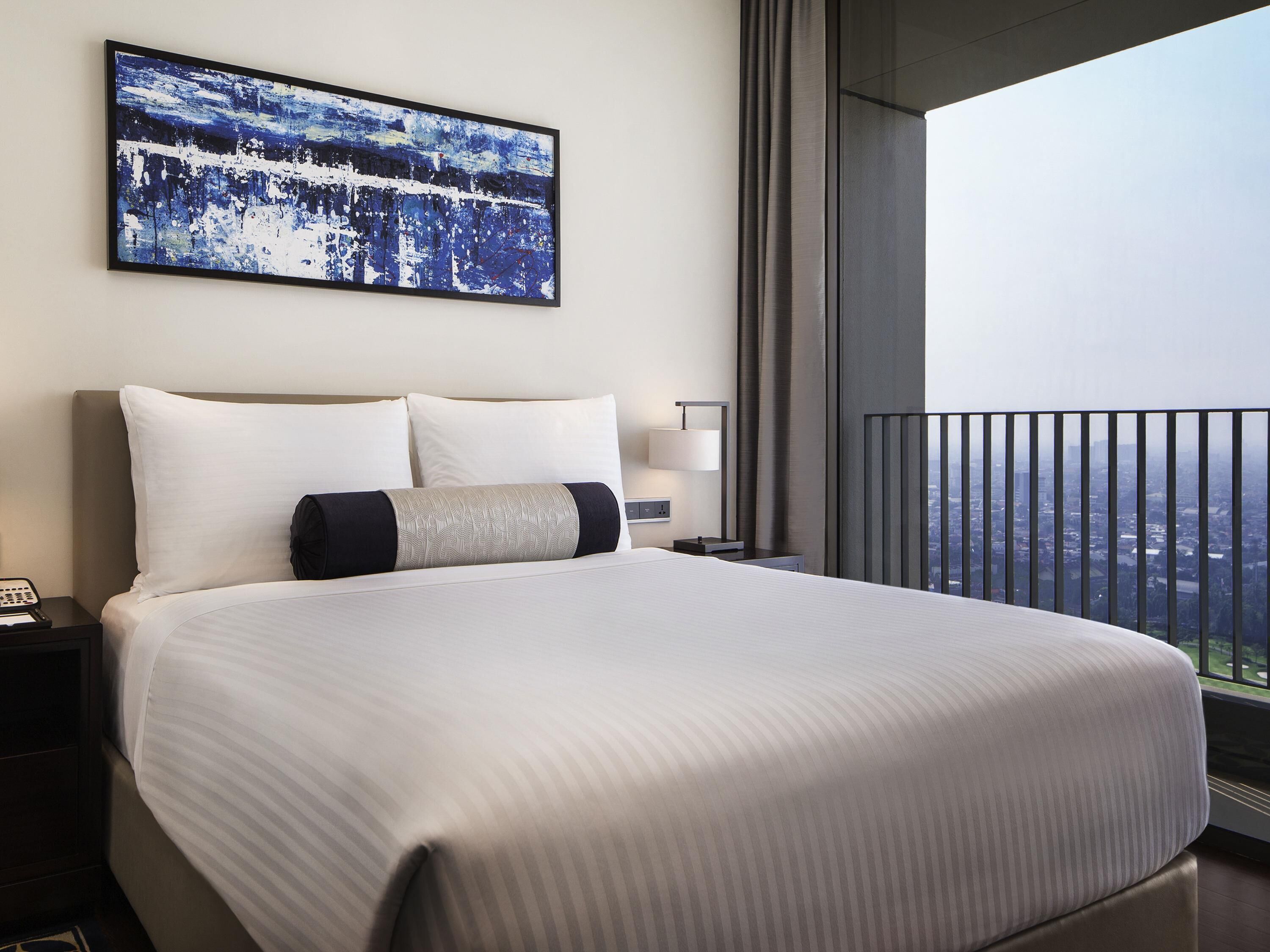 A hotel room with a large bed, a nightstand, a bedside lamp, a painting on the wall, and a balcony with a city view.