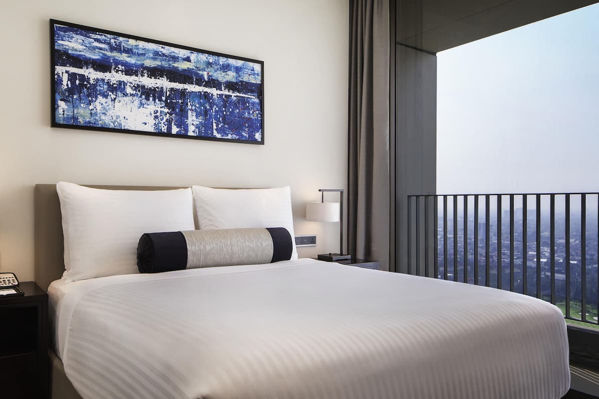 A hotel room with a large bed, a nightstand, a bedside lamp, a painting on the wall, and a balcony with a city view.