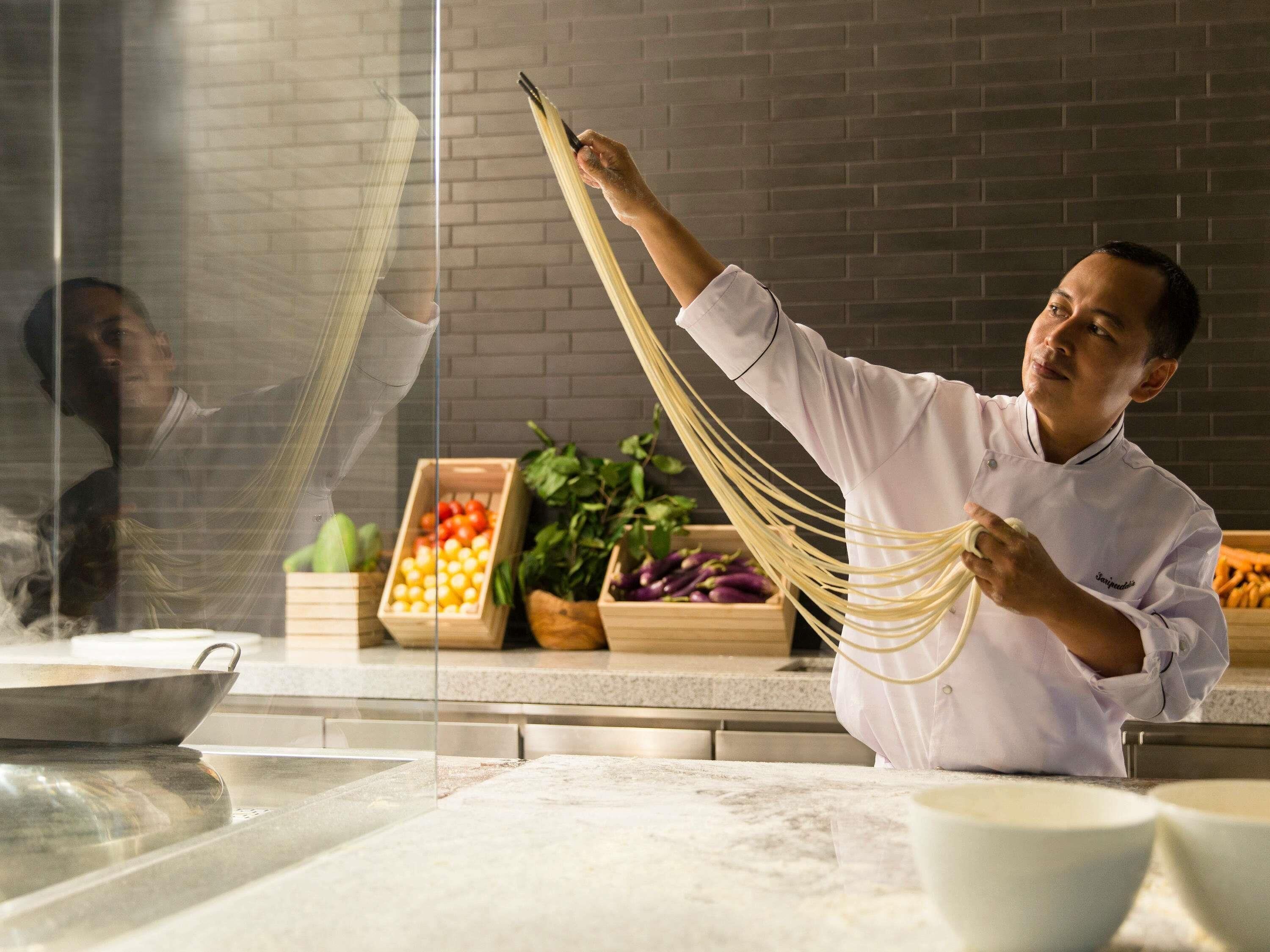 A chef preparing pasta in a kitchen with a brick wall and various vegetables in the background.