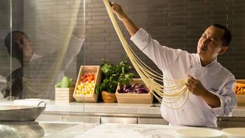 A chef preparing pasta in a kitchen with a brick wall and various vegetables in the background.