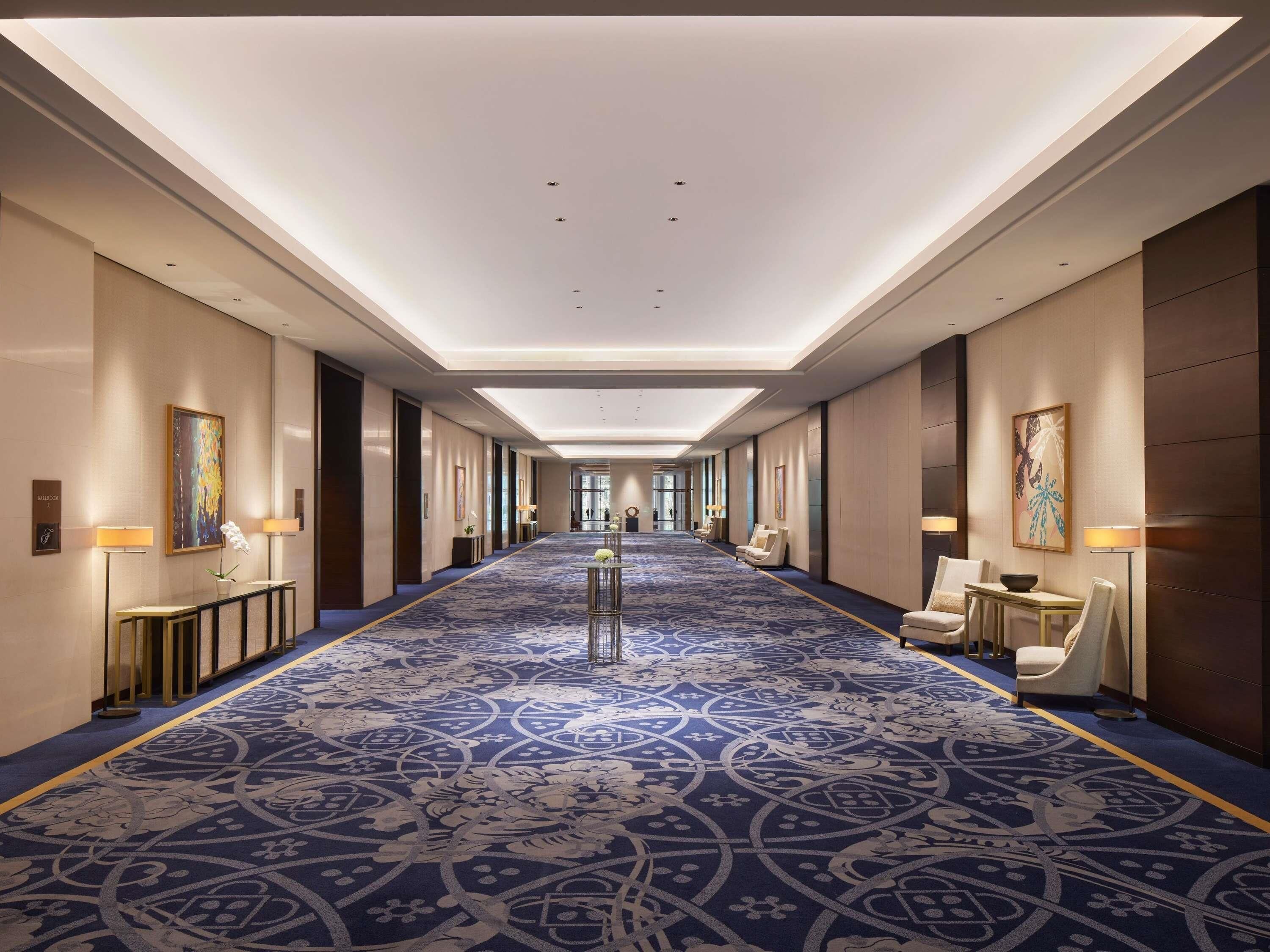 A long, narrow hotel corridor with a patterned carpet, wall-mounted lamps, and framed artwork.