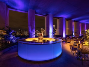 A rooftop bar with a circular seating area, surrounded by tall columns and palm trees, overlooking a city skyline at night. - Fairmont Jakarta (Jakarta)