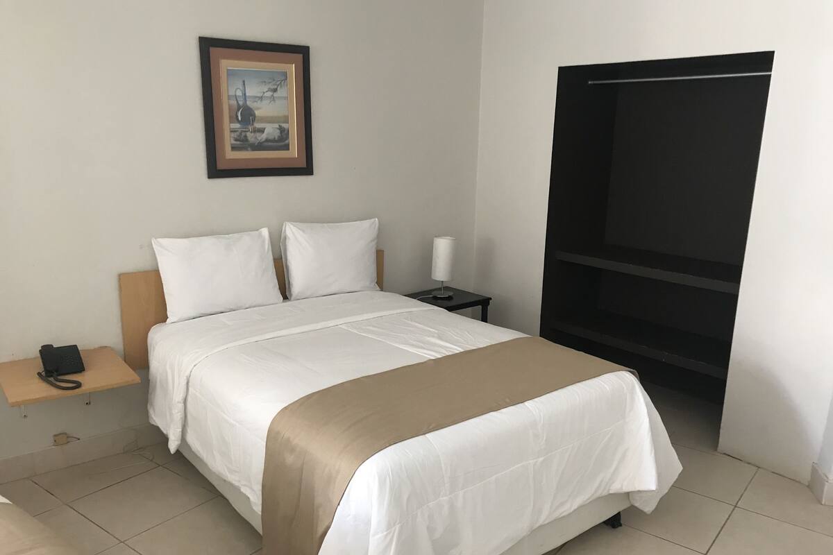 A hotel room with a bed, a bedside table with a phone, a lamp, and a painting on the wall.