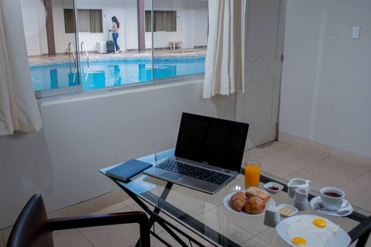 A room with a laptop on a table, breakfast items, and a view of a swimming pool.